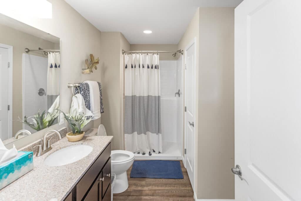 Modern bathroom with a shower, toilet, vanity with sink, mirror, and decorative towels. The floor is wood-style, and there is a blue bath mat in front of the shower.