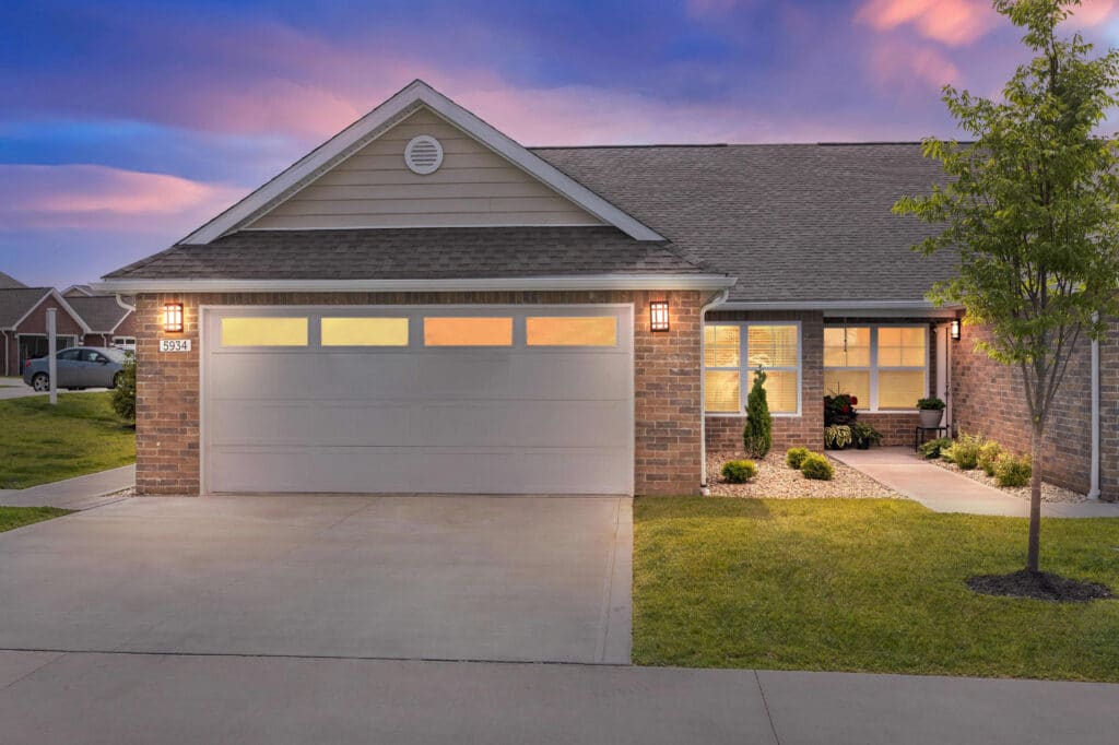 Single-story brick house with a two-car garage, lit porch lights, manicured lawn, and a tree, photographed at sunset.