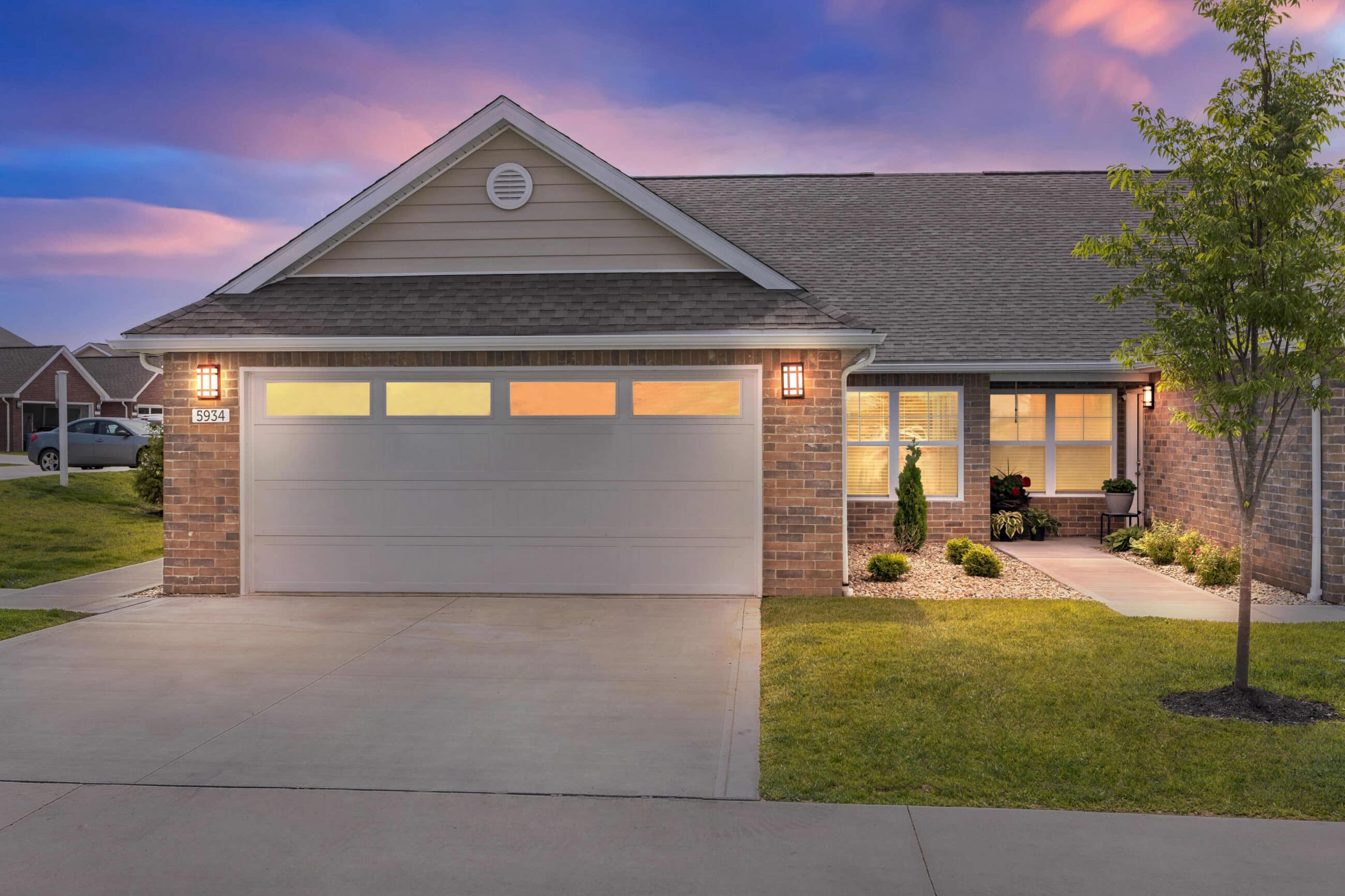 Single-story brick house with a two-car garage, lit porch lights, manicured lawn, and a tree, photographed at sunset.