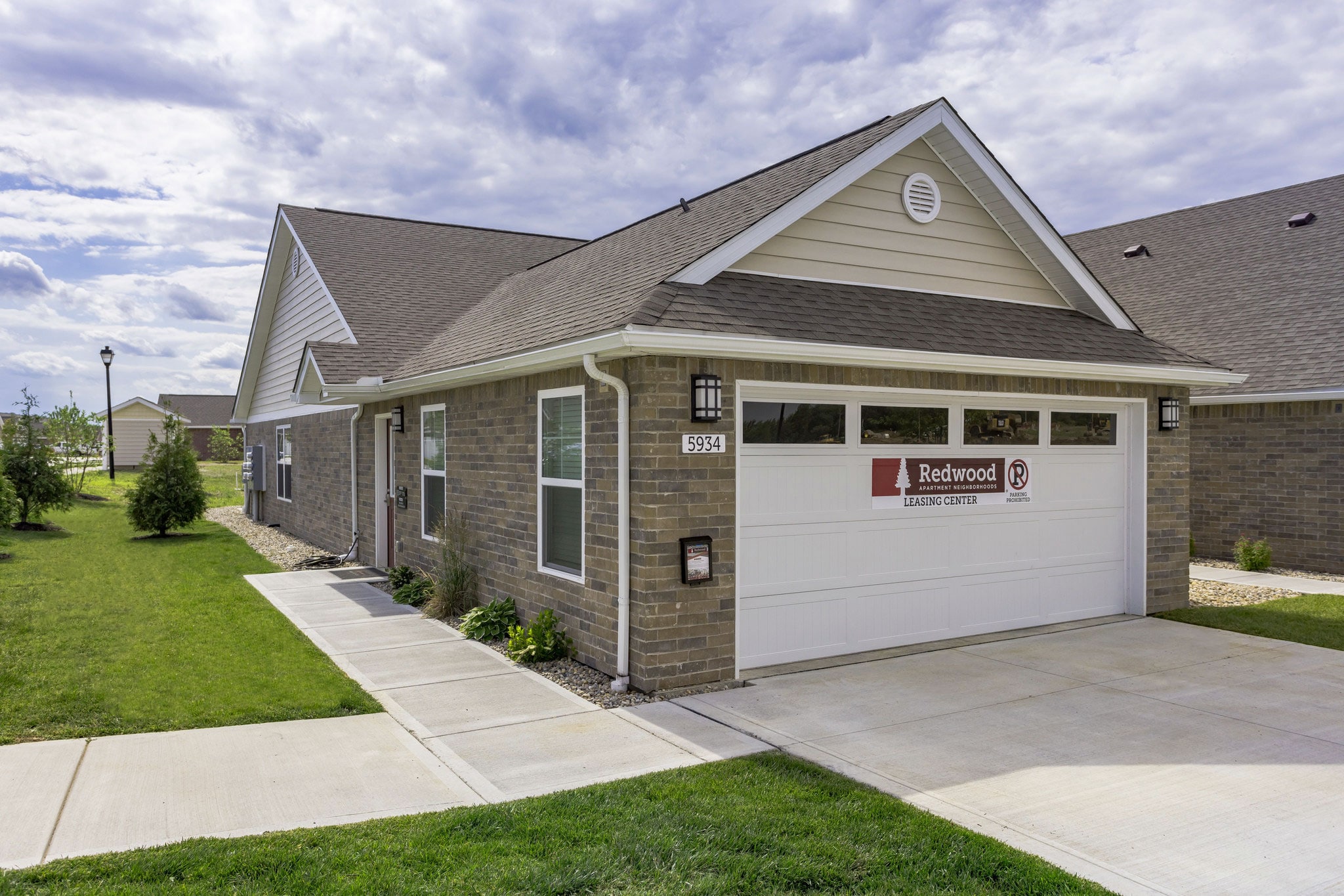 Single-story brick house with a gable roof, white garage door displaying a "Redwood" sign. The house is surrounded by a neat lawn and concrete driveway under a partly cloudy sky.