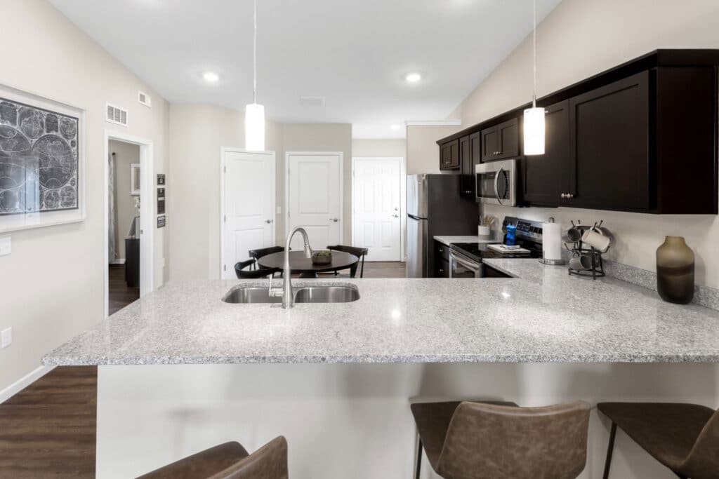 Modern kitchen with granite countertops, dark cabinets, stainless steel appliances, and a breakfast bar with three chairs; pendant lights hang above the island.