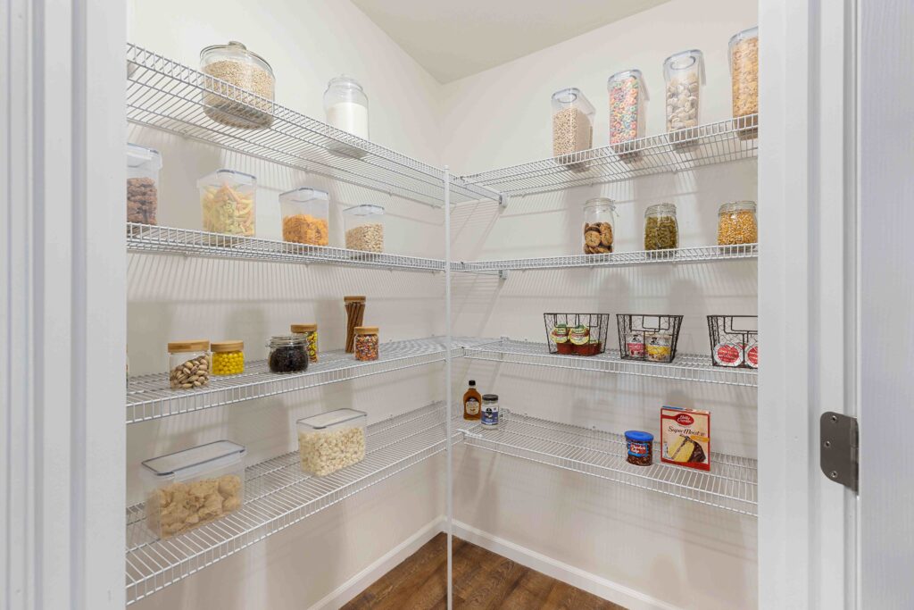 A neatly organized pantry with wire shelves holding clear containers of dry goods, glass jars, baskets of canned food, and a few bottles and boxes on wooden floors.