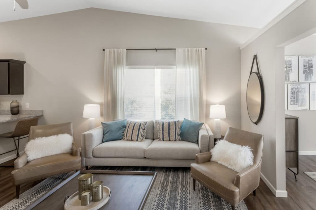 Modern living room with a beige sofa, two tan chairs with white pillows, a patterned rug, two side tables with lamps, and a round mirror on the wall.