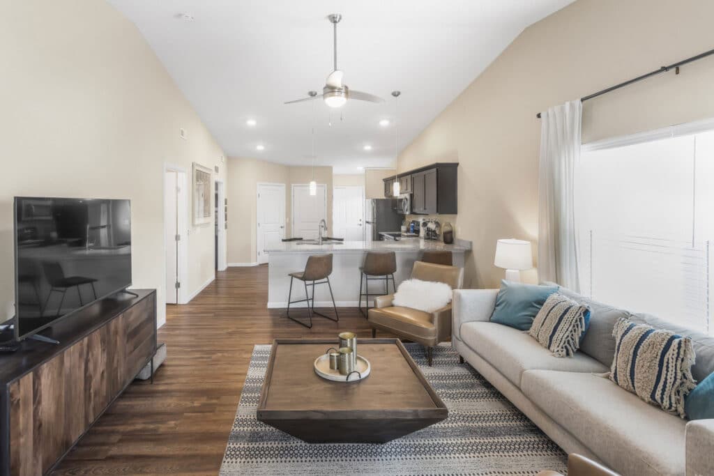 Modern open-concept living room and kitchen with neutral tones, a beige sofa, wooden coffee table, flat-screen TV, and barstools at a kitchen counter.
