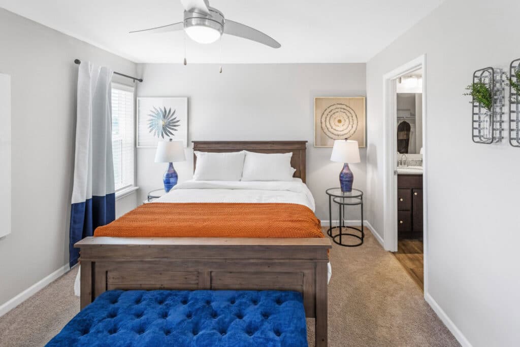 A neatly made bed with white bedding and an orange throw is centered in a bright bedroom with two lamps, wall art, and an open doorway leading to a bathroom.