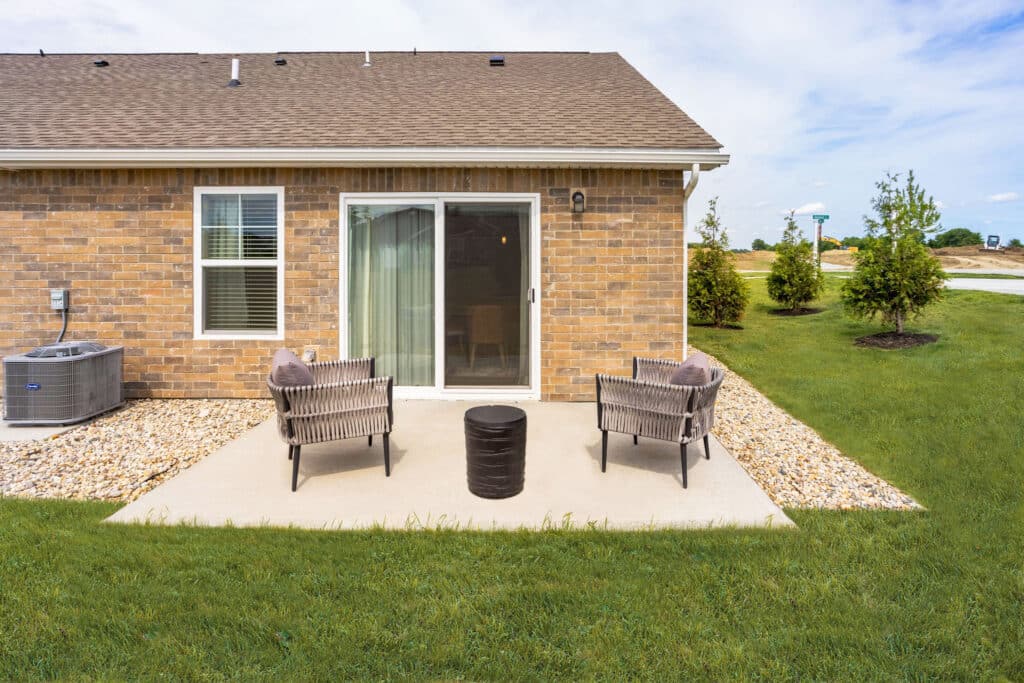 A small brick patio with two chairs and a round table sits outside a house with a sliding glass door and a grassy yard.