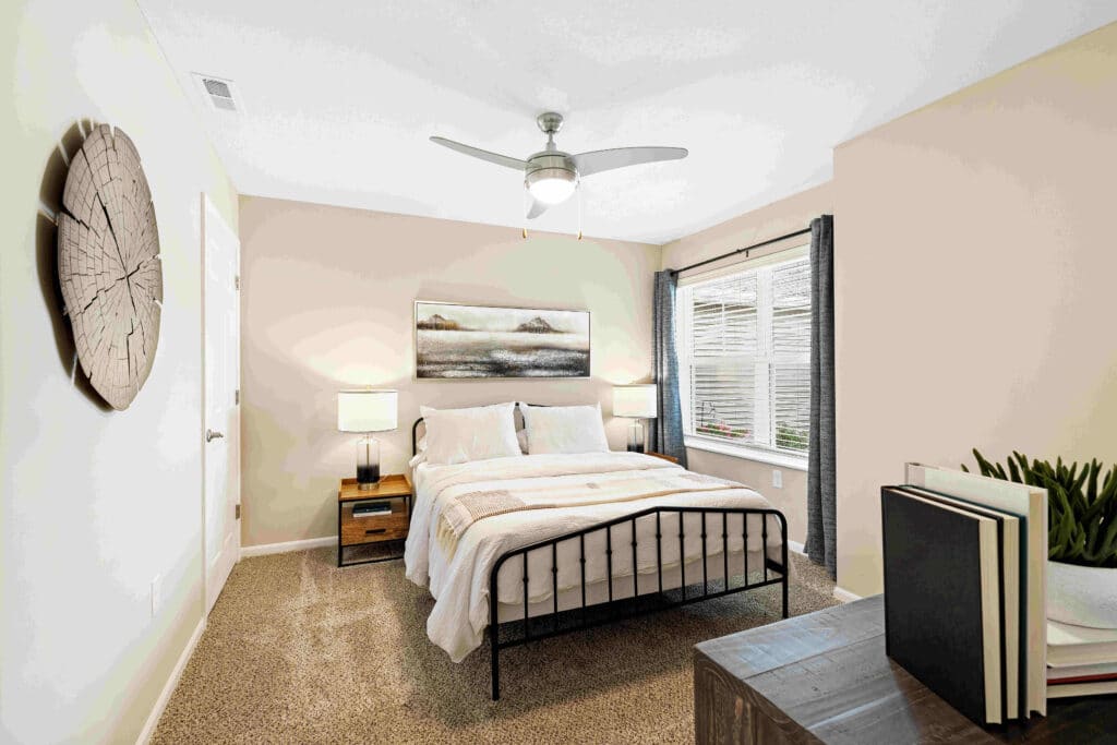 A neatly made bed with white bedding in a bright bedroom, featuring two nightstands, a ceiling fan, large window, and wall art above the headboard.