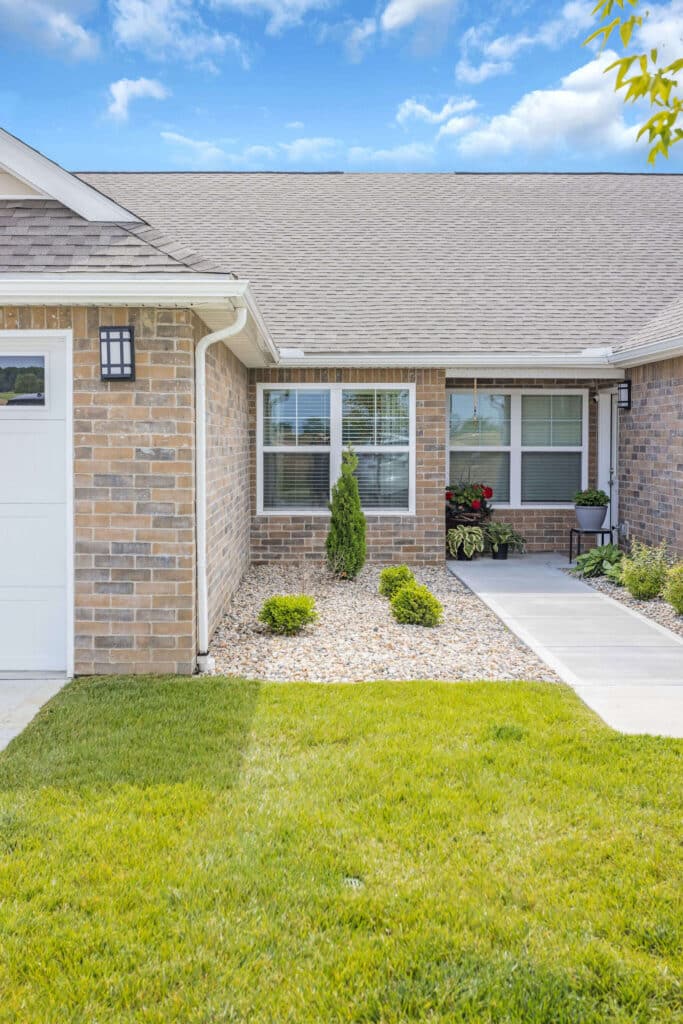 Single-story brick house with a two-window front, a small garden with rocks and shrubs, and a concrete walkway leading to the entrance.