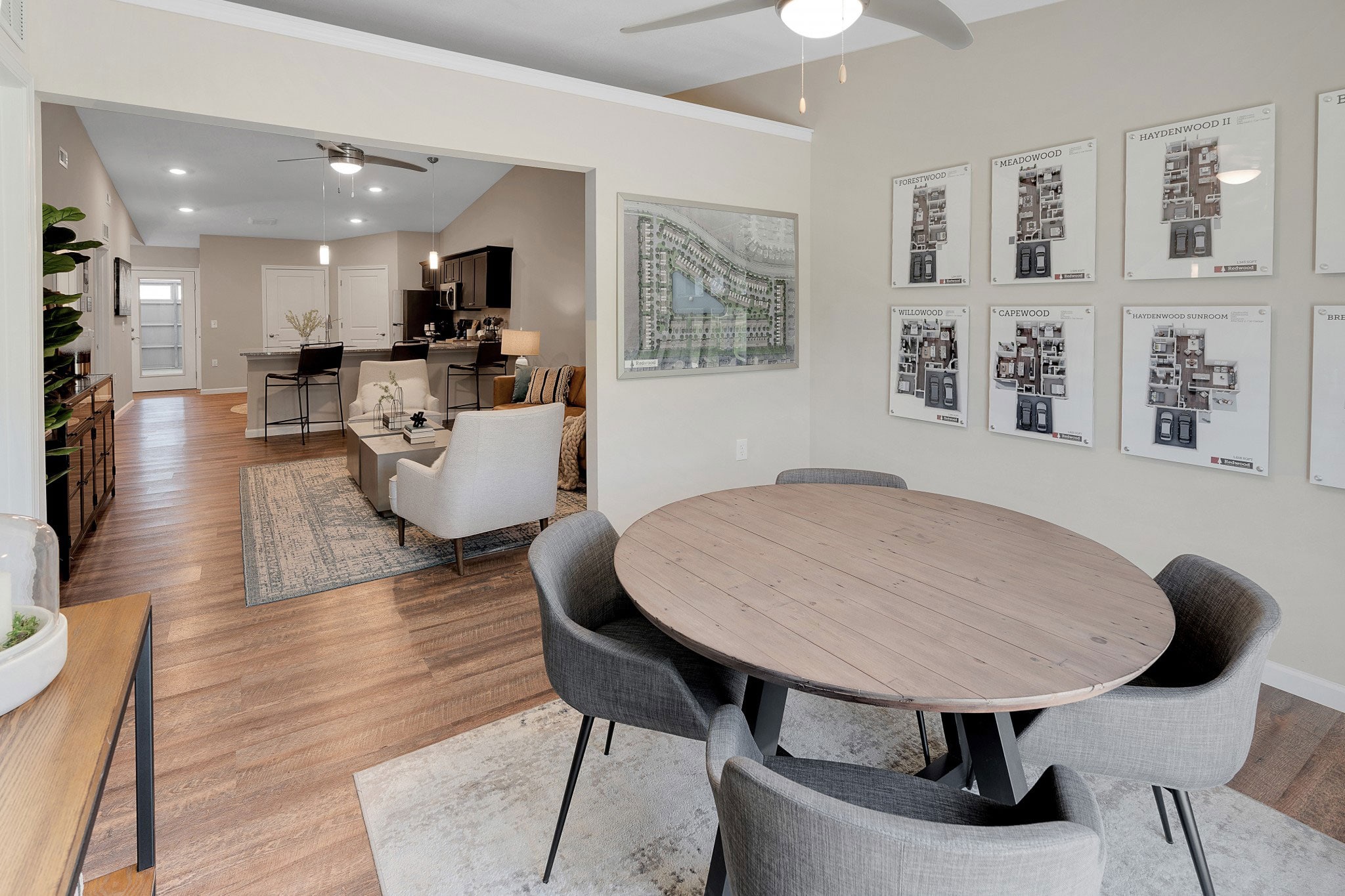 Dining area with a round wooden table and four gray chairs. Open concept leads to a living room with two beige armchairs and a kitchen. Floor plans are displayed on the wall.