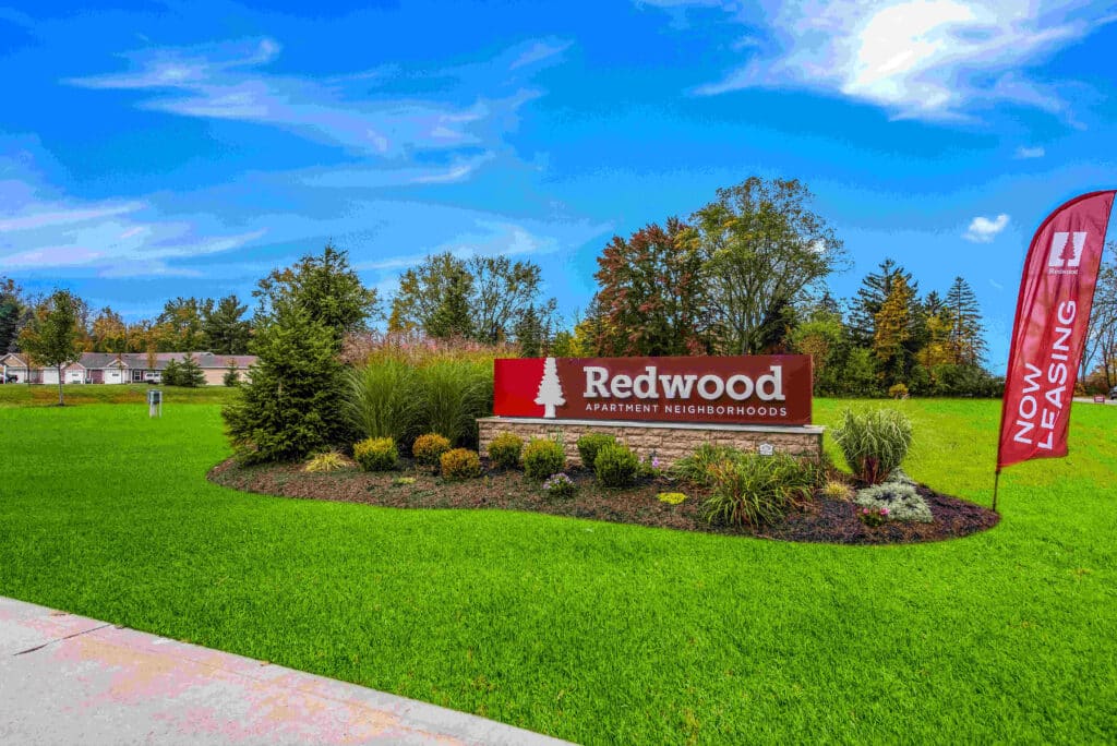 Redwood Apartment Neighborhoods sign on a landscaped area with green grass and shrubs, under a blue sky. A "Now Leasing" flag stands beside it. Trees and buildings are visible in the background.