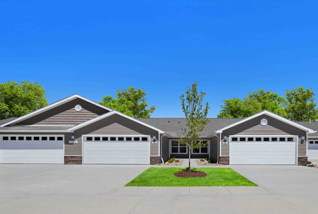 Row of identical single-story homes with peaked roofs and two-car garages, separated by driveways. Small tree and grass patch in front, clear blue sky above.