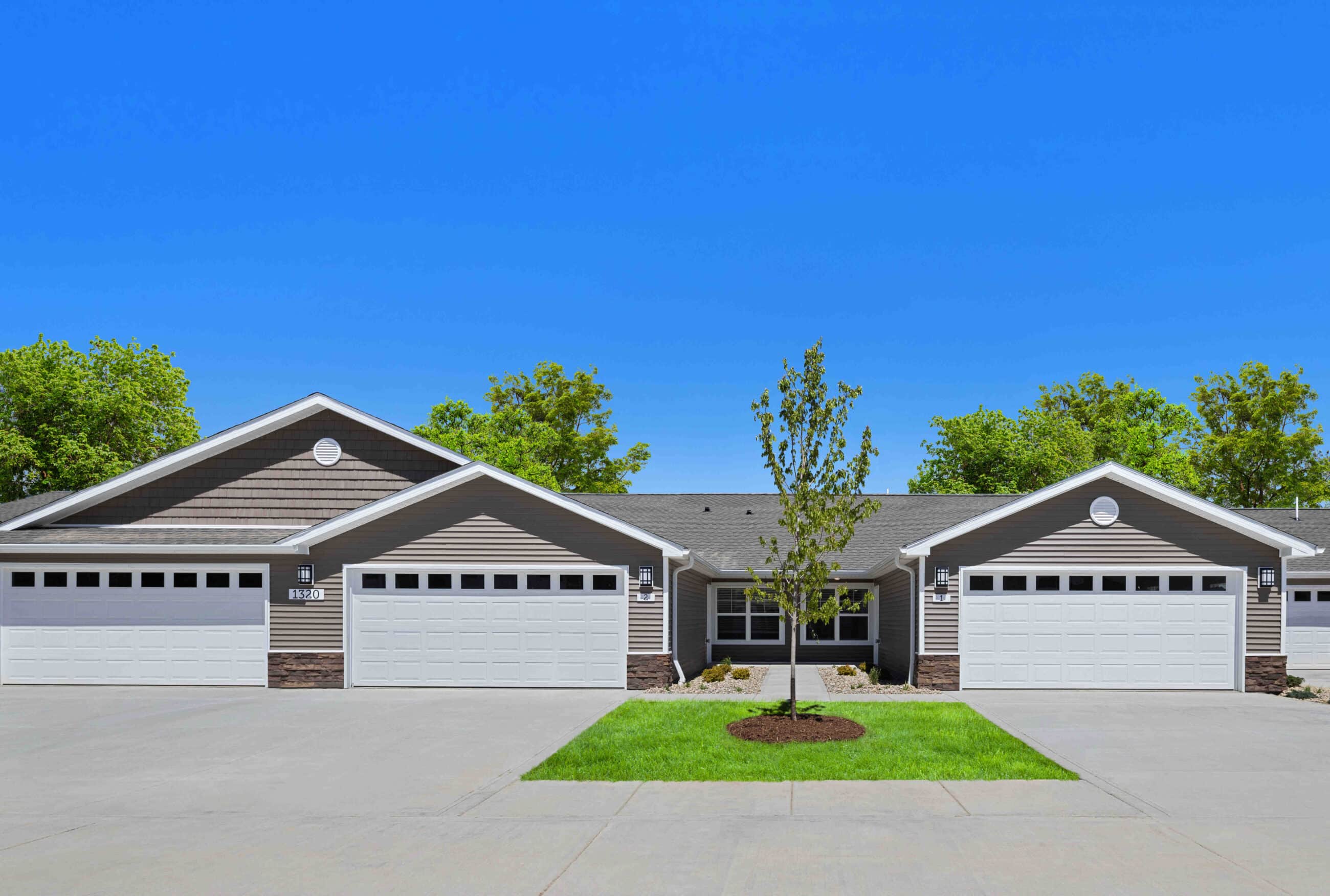 Row of identical single-story homes with peaked roofs and two-car garages, separated by driveways. Small tree and grass patch in front, clear blue sky above.