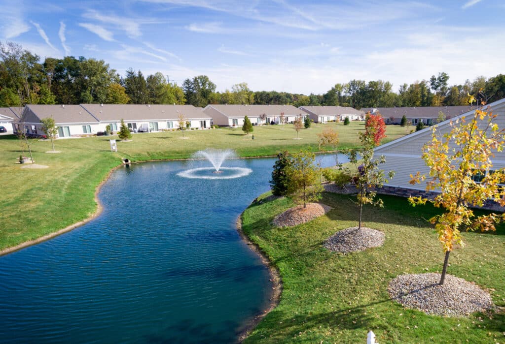 A small pond with a central fountain is surrounded by grass and trees. Single-story houses are in the background under a blue sky with wispy clouds.