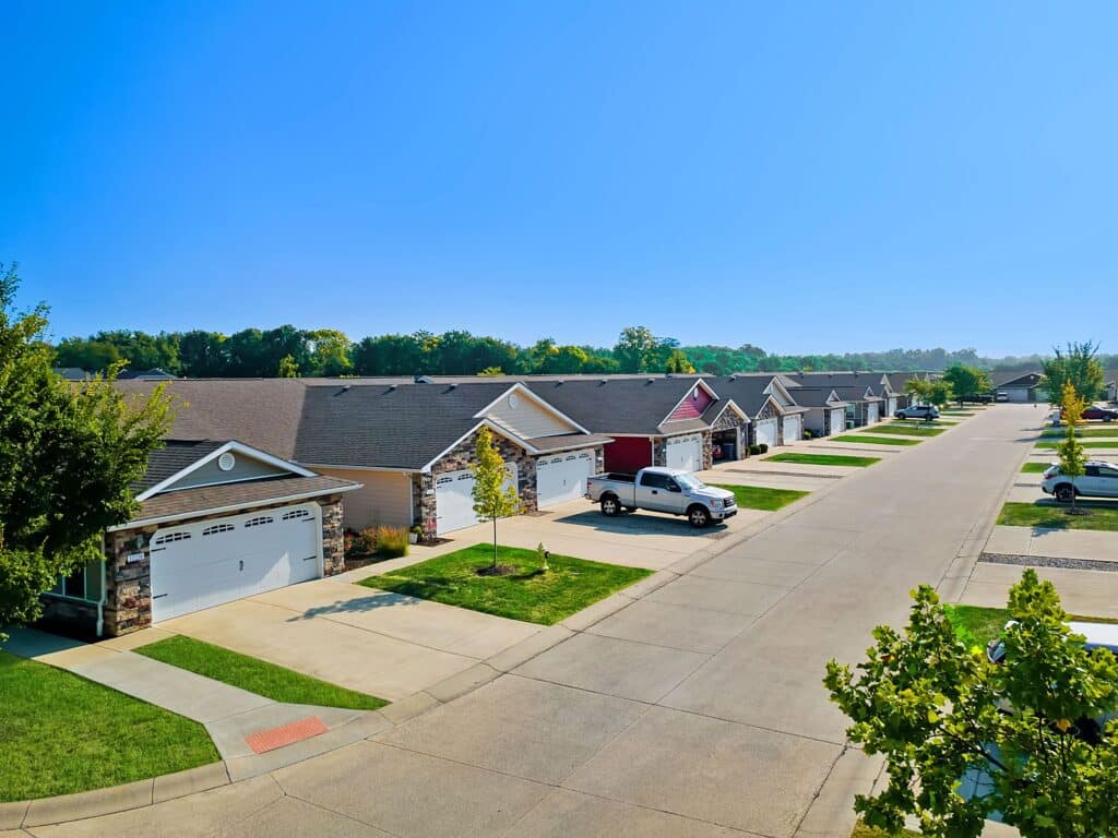 A suburban street with single-story houses, each with a driveway and garage, neatly trimmed lawns, and several parked vehicles on a clear, sunny day.