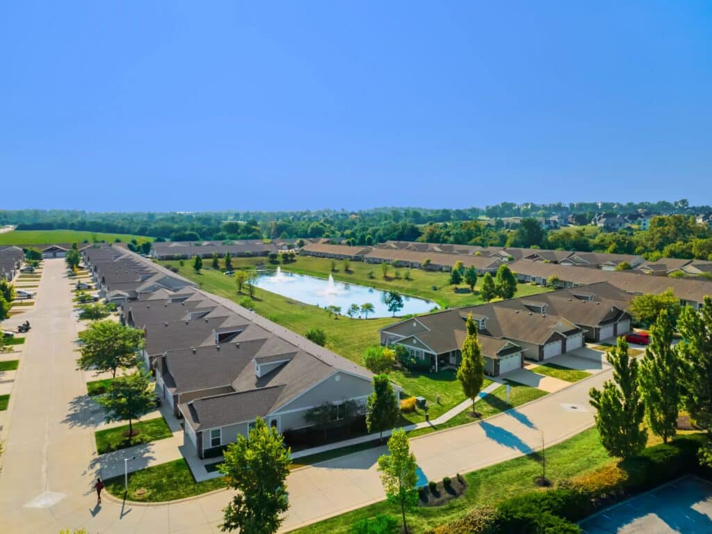 Aerial view of a suburban neighborhood with rows of houses, green lawns, trees, and a central pond under a clear blue sky.