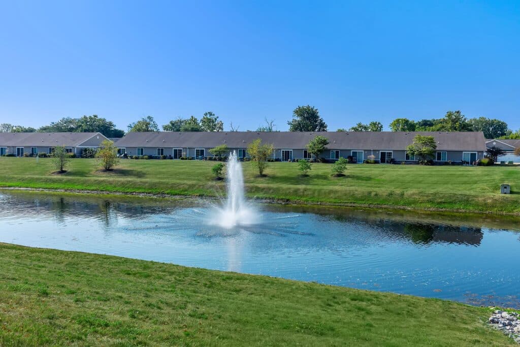 A row of single-story homes faces a small pond with a central water fountain, surrounded by green grass and trees under a clear blue sky.