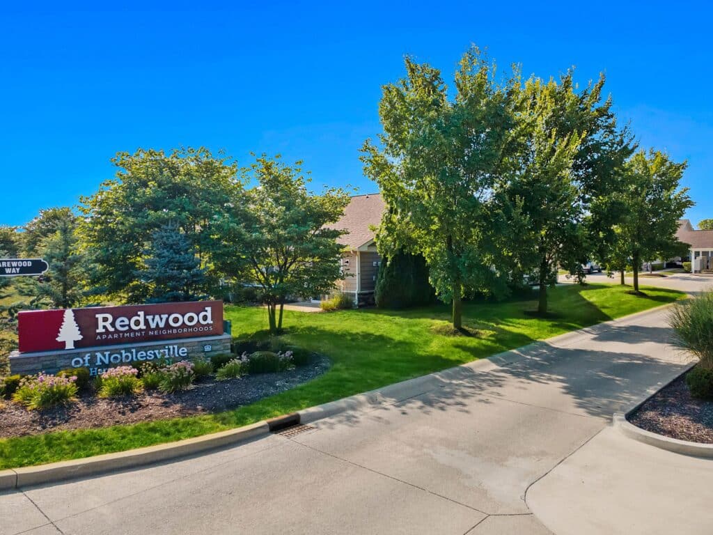 Entrance to Redwood Apartment Neighborhoods of Noblesville, featuring a large red sign, trees, and a paved driveway on a clear day.
