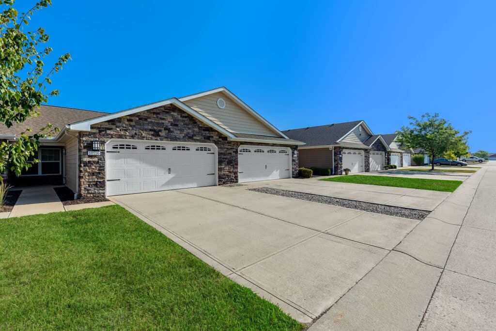 Row of modern suburban homes with attached garages, stone and siding exteriors, concrete driveways, manicured lawns, and clear blue sky.