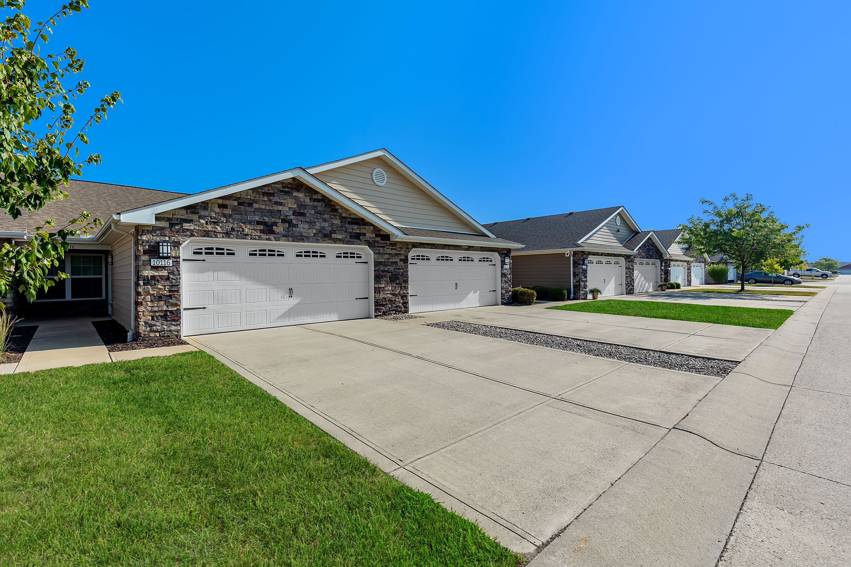 Row of modern suburban homes with attached garages, stone and siding exteriors, concrete driveways, manicured lawns, and clear blue sky.