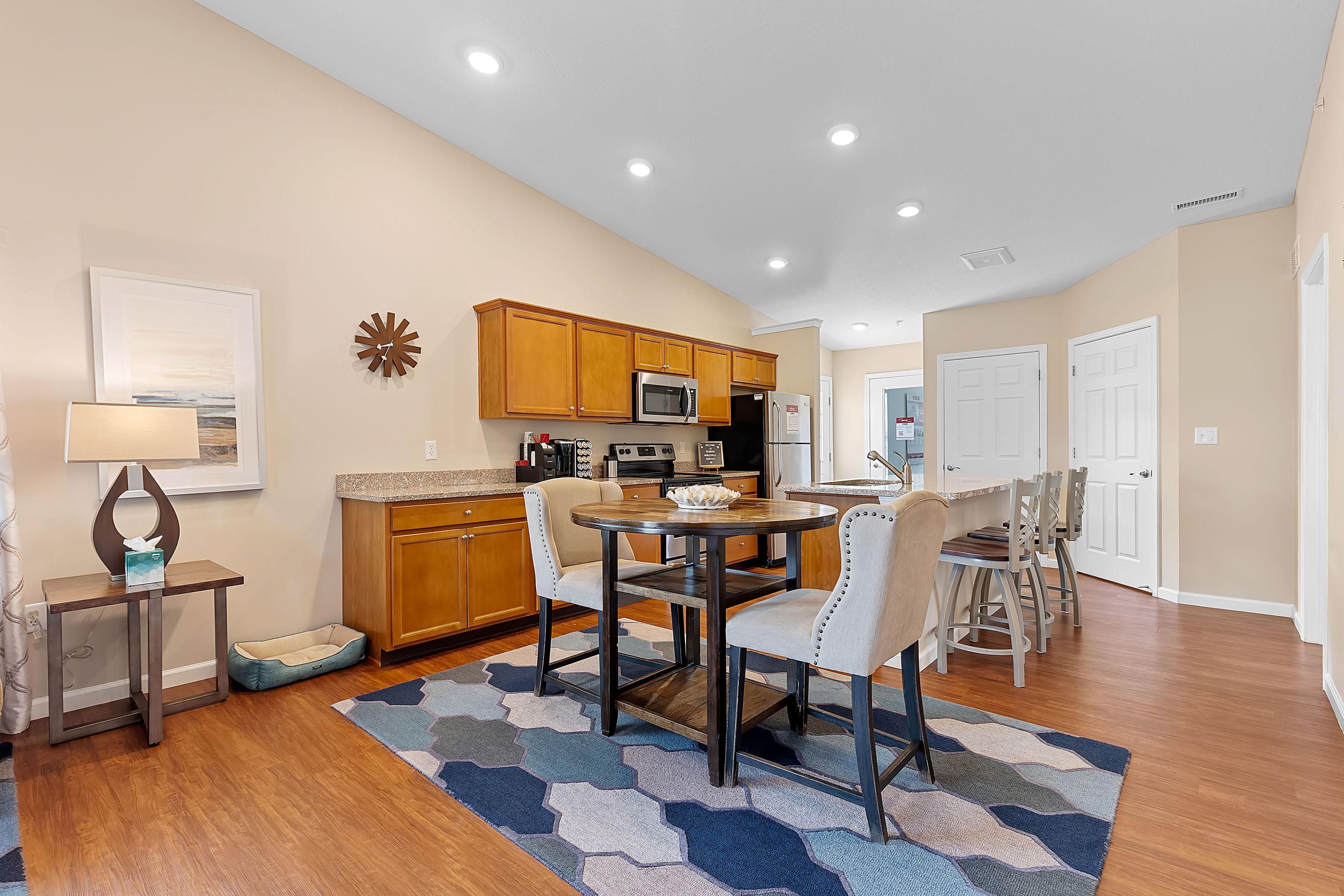 A modern kitchen and dining area with wooden cabinets, stainless steel appliances, a round dining table, upholstered chairs, and a patterned rug on hardwood flooring.