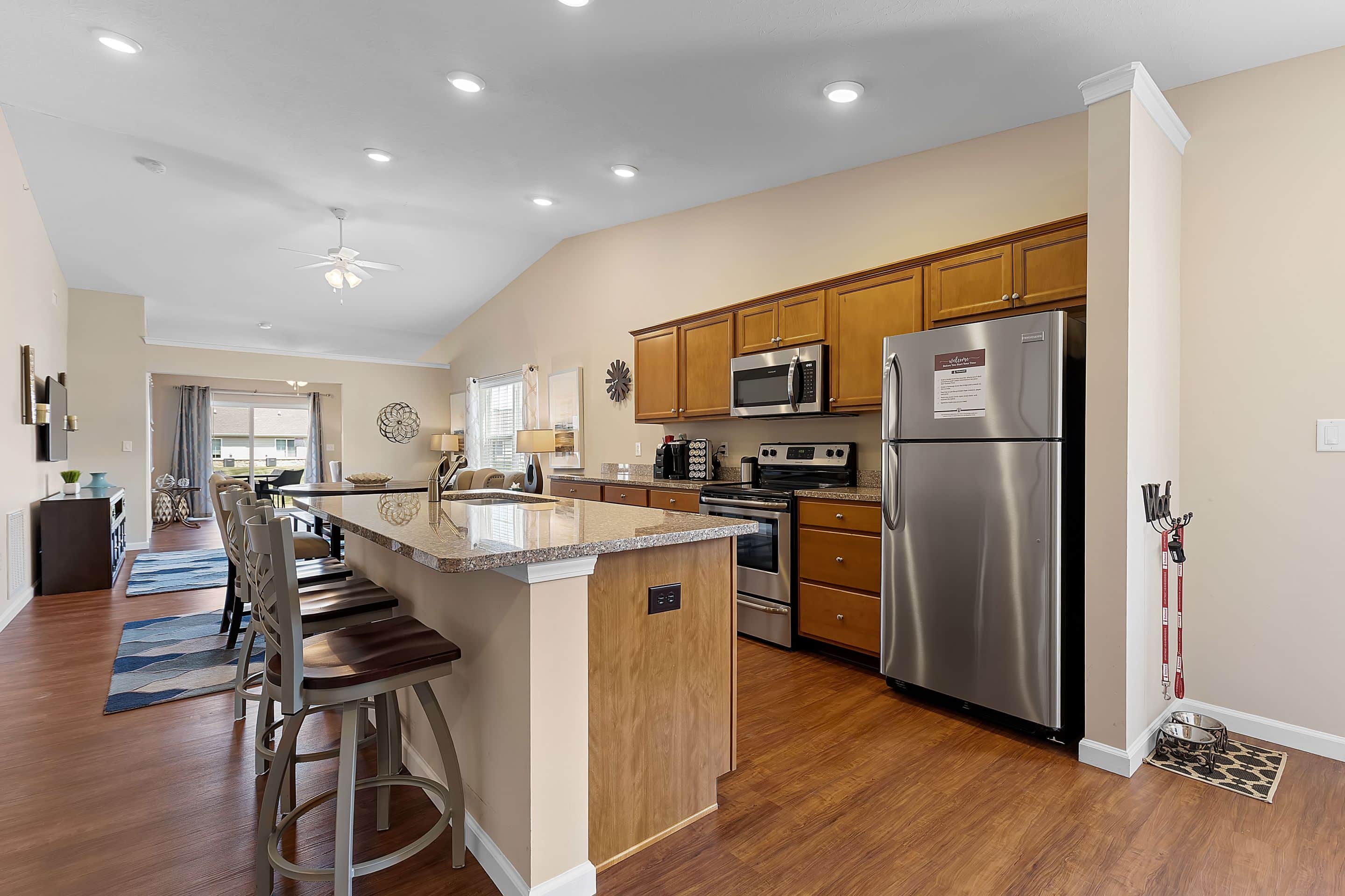 Modern kitchen with stainless steel appliances, wooden cabinets, and granite countertops, connected to a dining area and living space with wooden flooring and neutral walls.