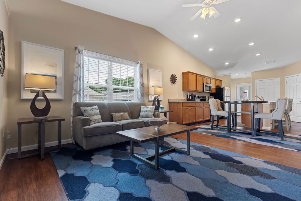 Open living space with a couch, coffee table, dining area, and kitchen. Blue geometric rug covers hardwood floor. Neutral walls, large window, ceiling fan, and recessed lighting.