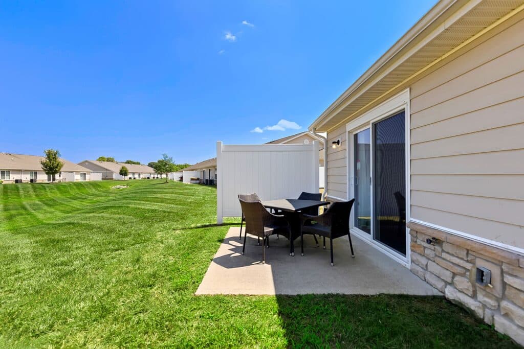 Patio area with a table and four chairs on a concrete slab beside a beige house, overlooking a large grassy lawn under a clear blue sky.