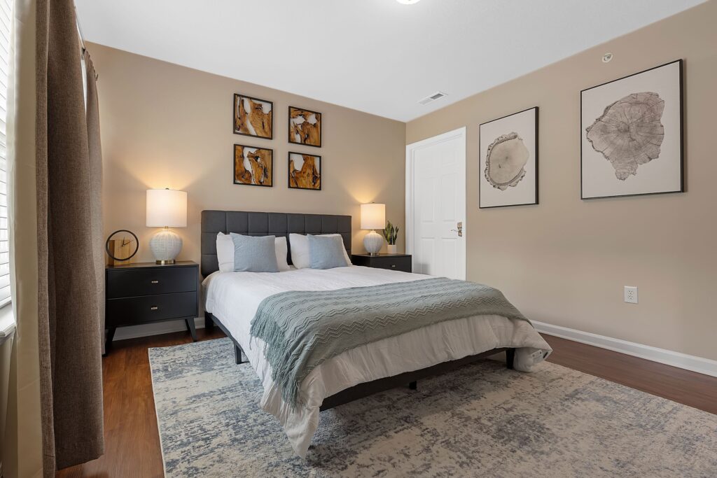 A neatly made bed with blue and white bedding in a modern bedroom, featuring two bedside tables, lamps, wall art, and a large window with curtains.