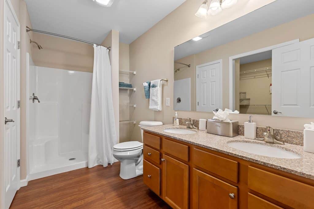 Bathroom with double sink vanity, granite countertop, wood cabinets, toilet, walk-in shower with white curtain, towel rack, and large mirror above the sinks.