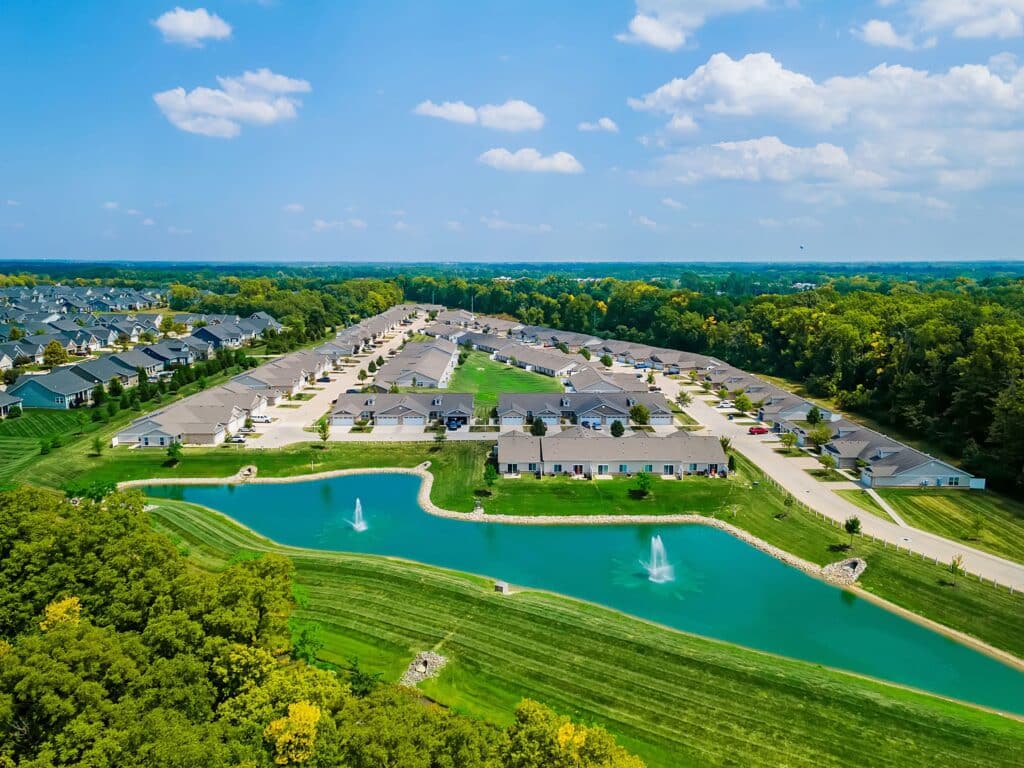 Aerial view of a suburban neighborhood with rows of houses, a pond with two fountains in the foreground, and trees surrounding the area under a partly cloudy sky.
