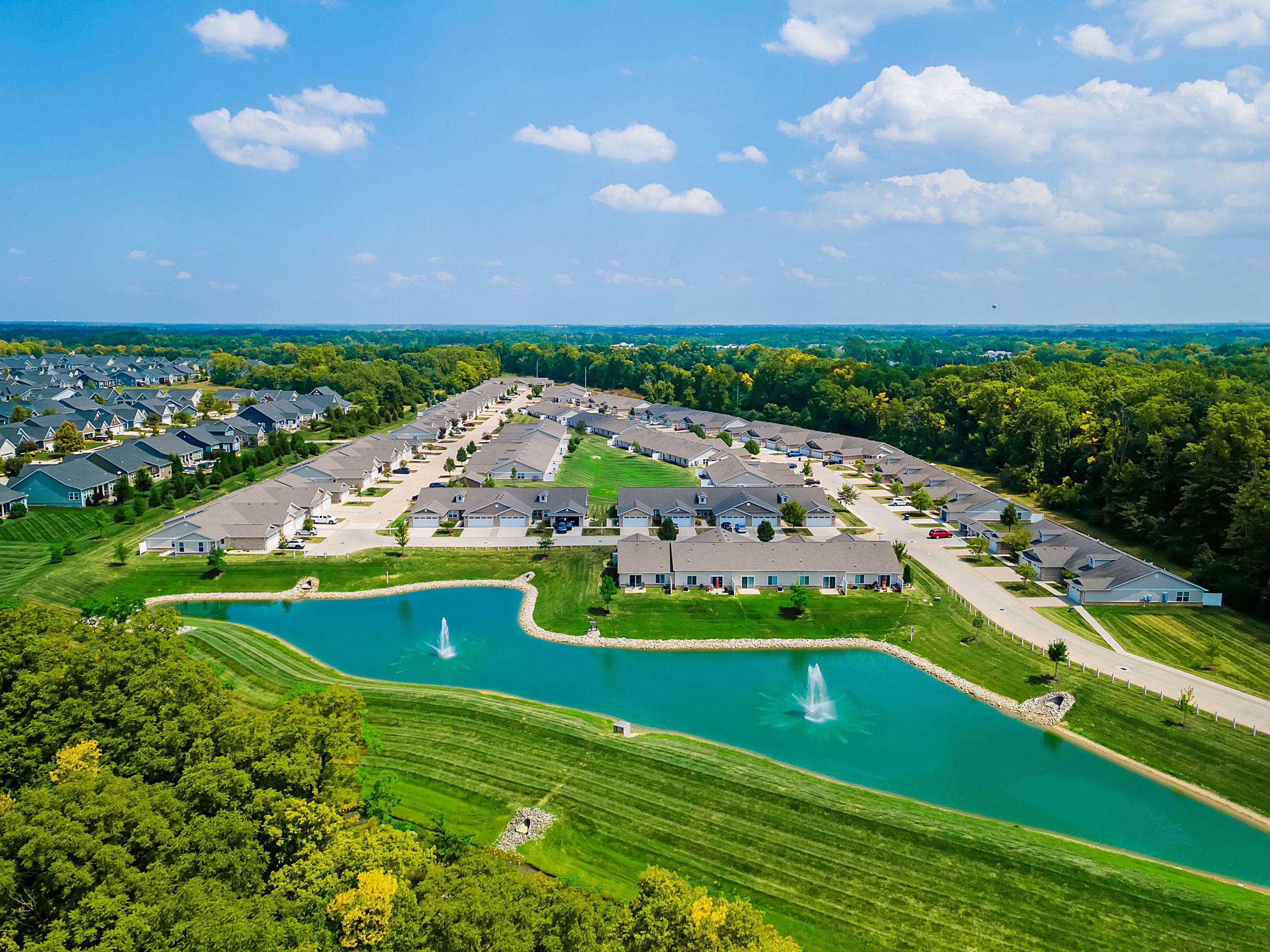 Aerial view of a suburban neighborhood with rows of houses, a pond with two fountains in the foreground, and trees surrounding the area under a partly cloudy sky.