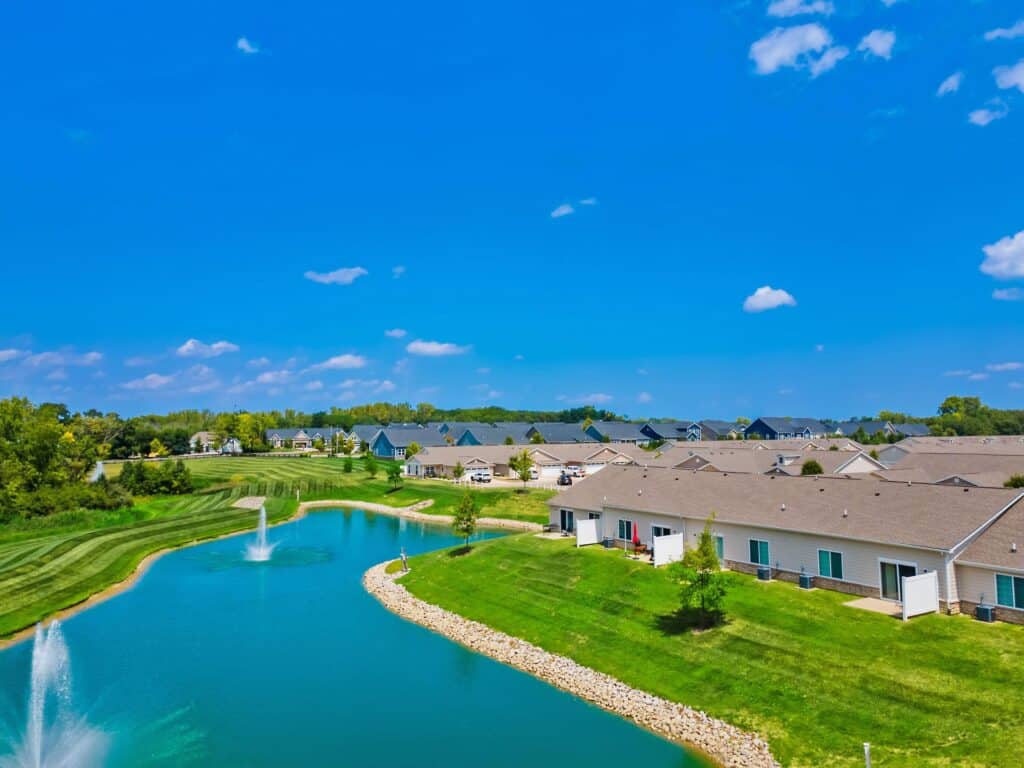 A suburban neighborhood with single-story houses, a pond with fountains, green lawns, and trees under a bright blue sky with scattered clouds.