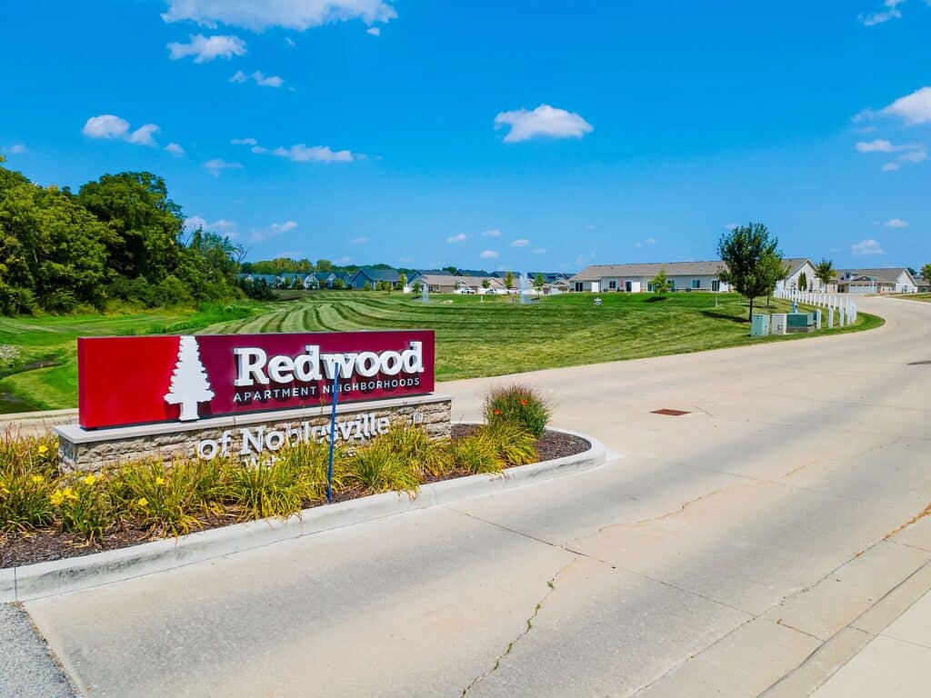 Redwood Apartment Neighborhoods sign at an entrance with landscaped plants, wide road, and residential houses in the background under a clear blue sky.