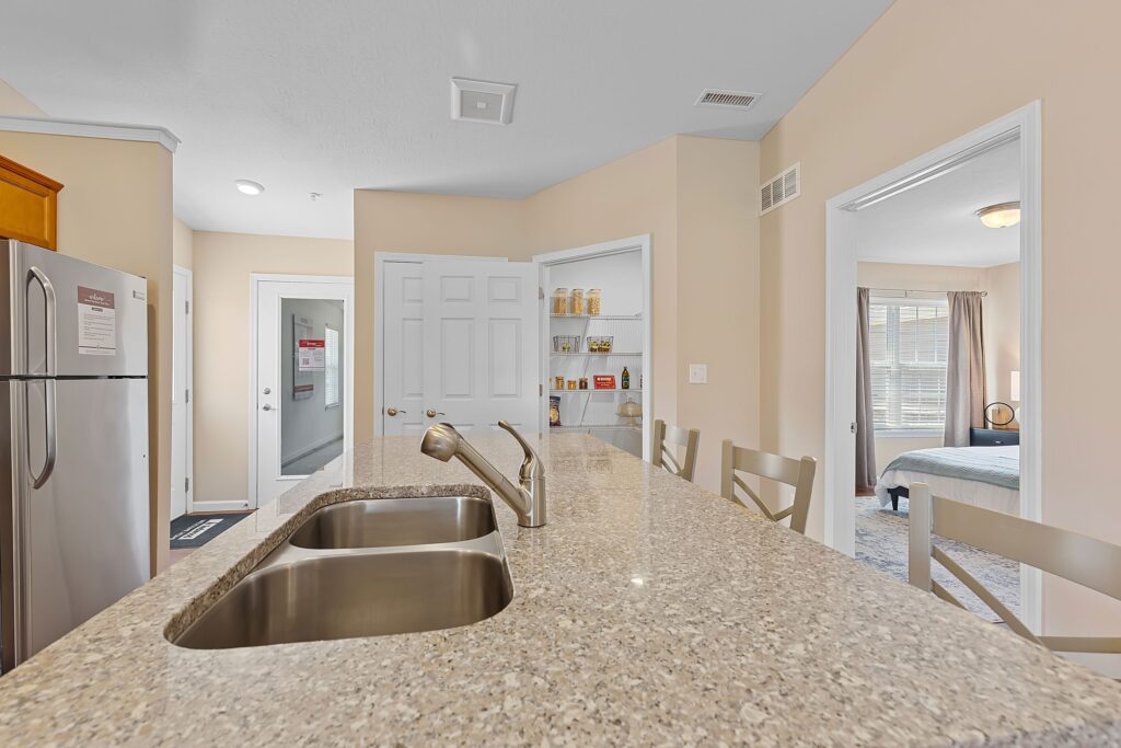 Modern kitchen with granite countertop, double sink, and stainless steel fridge; chairs line the island, and a pantry and bedroom are visible in the background.