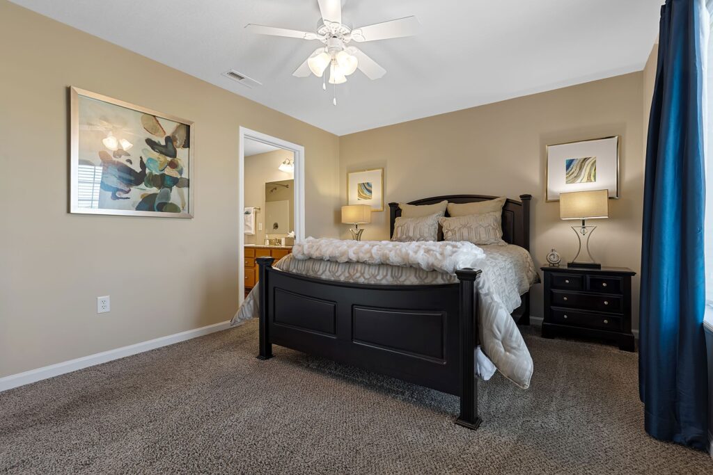 A bedroom with a dark wooden bed, two nightstands with lamps, framed artwork on beige walls, carpeted floor, and a ceiling fan. Doorway leads to an adjacent bathroom.