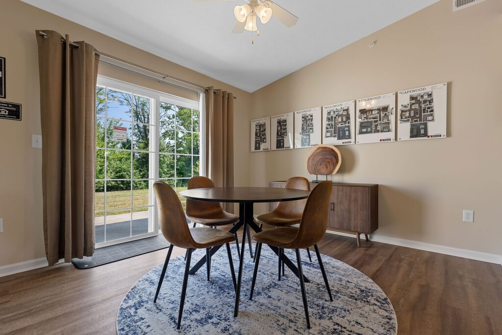 A modern dining area with a round table and four chairs on a blue and white rug, next to sliding glass doors and a sideboard with framed wall art above it.