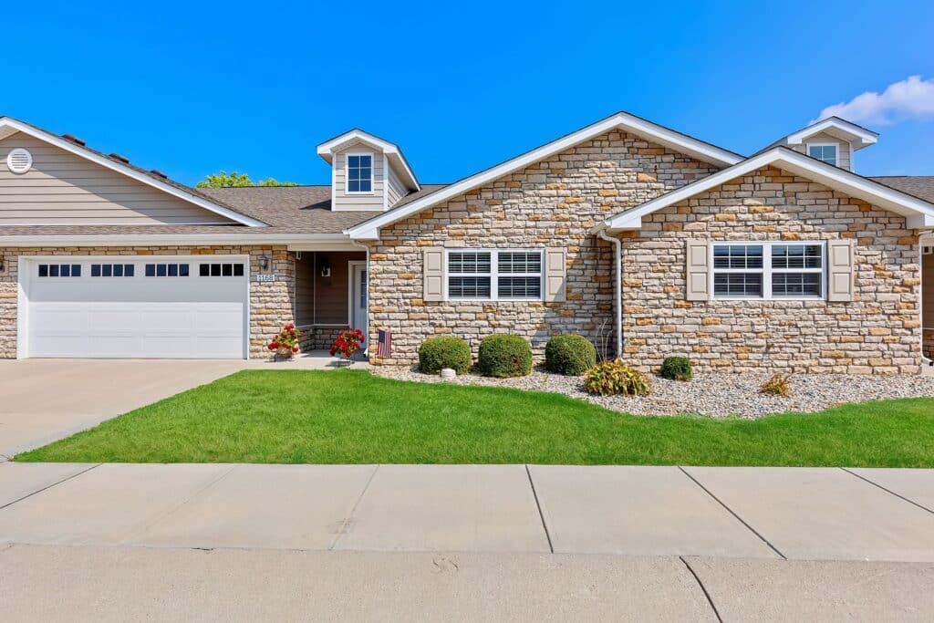 Single-story house with stone facade, white trim, attached two-car garage, green lawn, shrubs, and a concrete driveway under a clear blue sky.