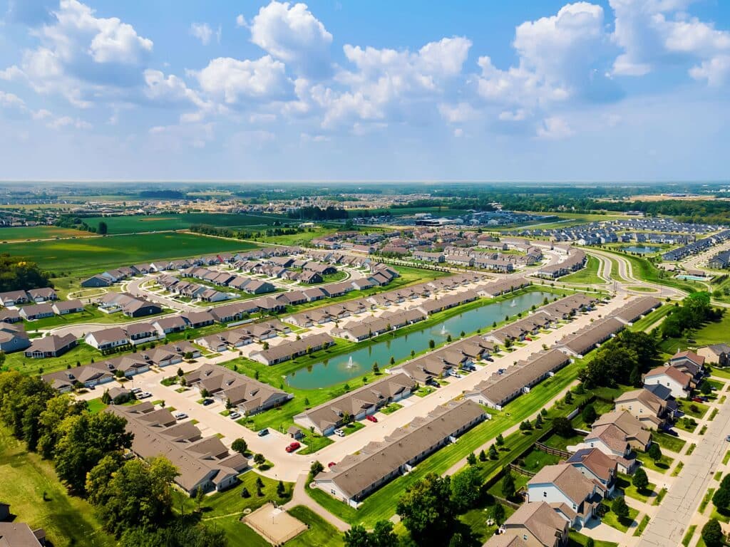 Aerial view of a suburban neighborhood with rows of houses, a central pond, green lawns, and surrounding roads under a partly cloudy sky.