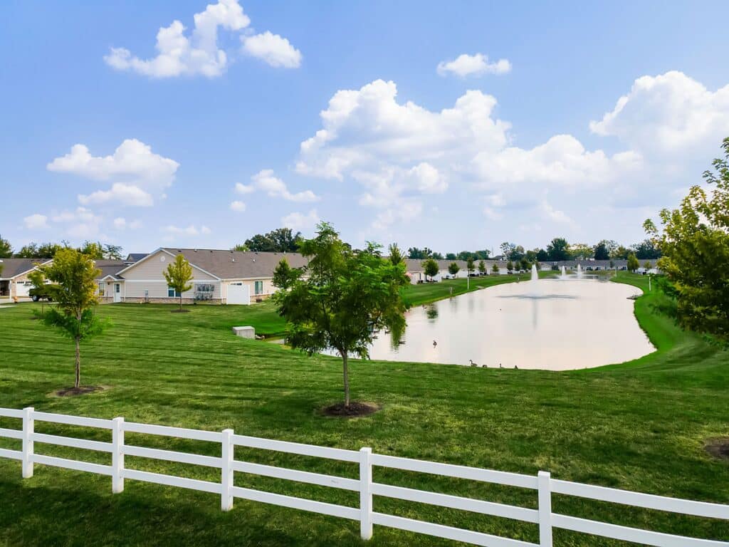 A suburban neighborhood with single-story houses, a white fence, green lawns, young trees, and a small pond under a partly cloudy sky.