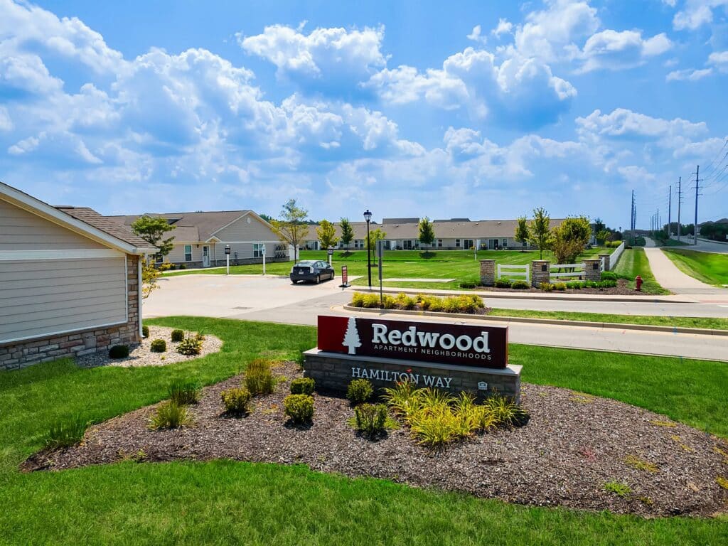 A landscaped entrance sign reading "Redwood Apartment Neighborhoods Hamilton Way" sits in front of single-story residential buildings on a sunny day.