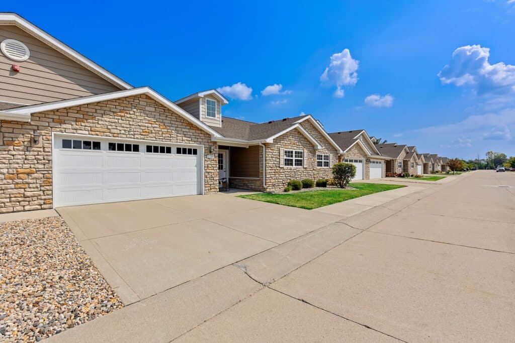 Row of modern suburban houses with stone and siding exteriors, attached garages, driveways, and well-maintained lawns on a clear, sunny day.