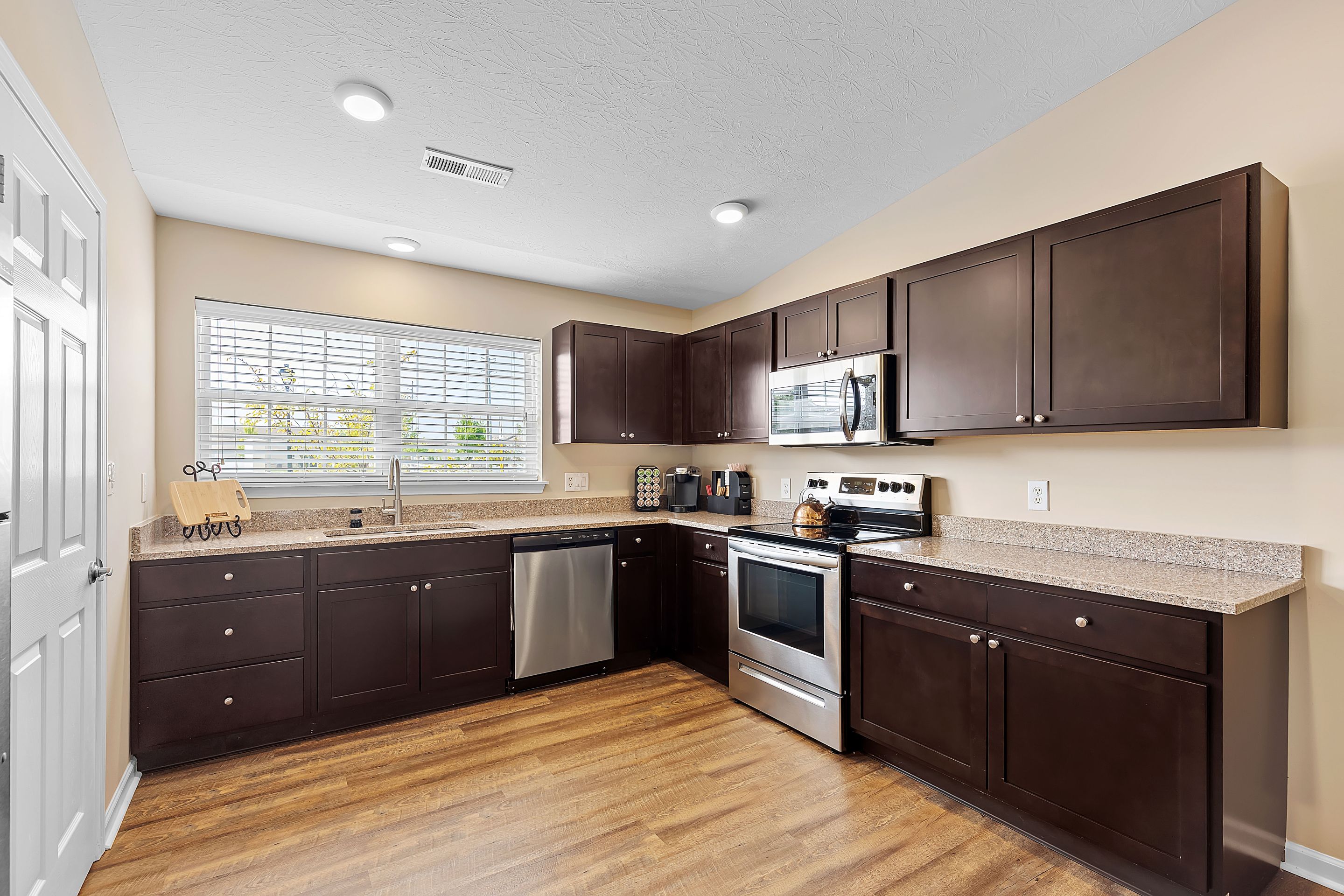 Modern kitchen with dark wood cabinets, stainless steel appliances, granite countertops, and wood flooring, illuminated by natural light from a large window.