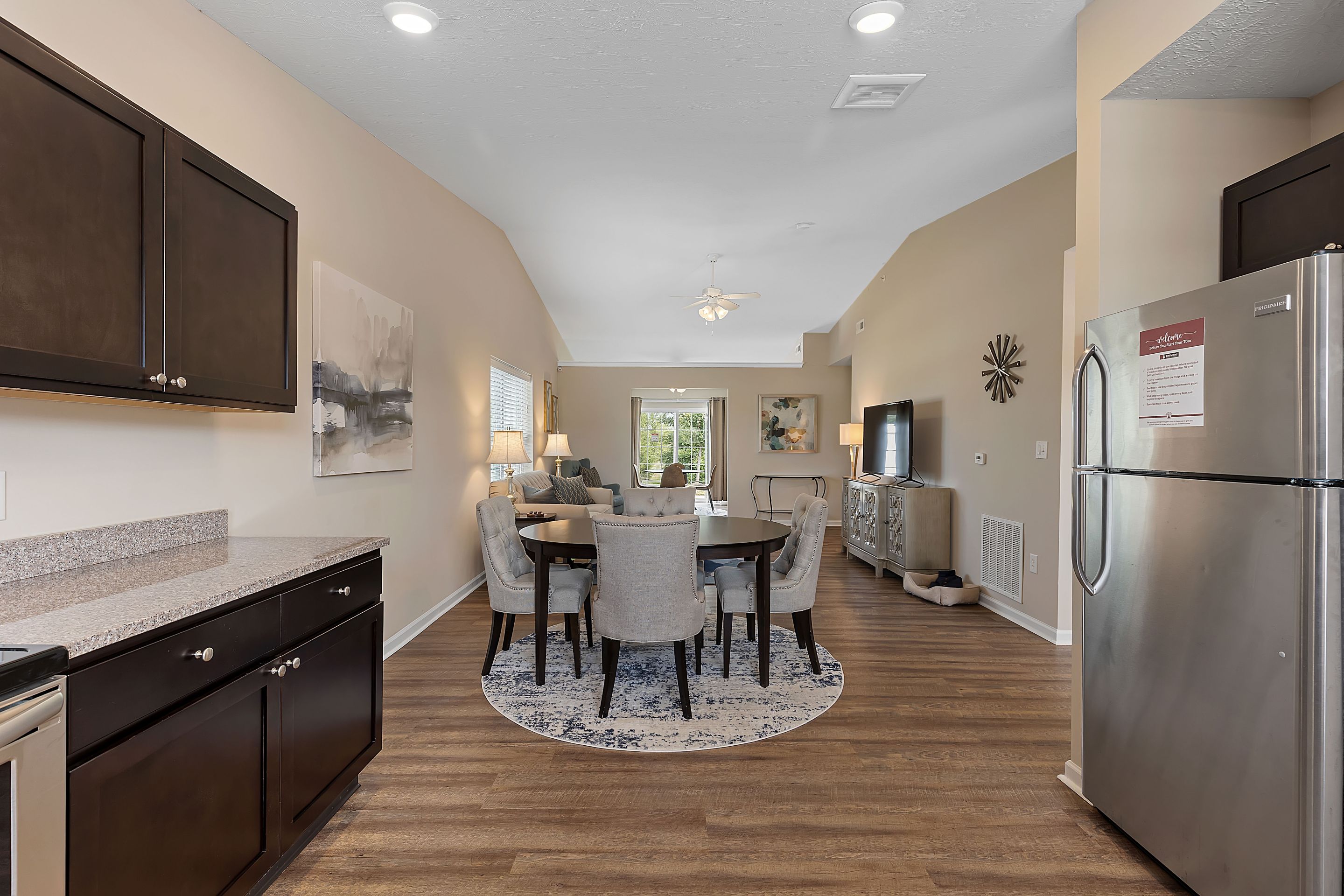 Open-concept kitchen and dining area with dark cabinets, round dining table and chairs, hardwood floors, and a living room with TV and sofa visible in the background.