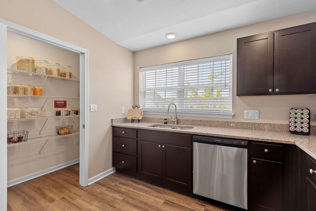 Modern kitchen with dark cabinets, stainless steel dishwasher, and a granite countertop next to a pantry with wire shelving holding jars of food items.