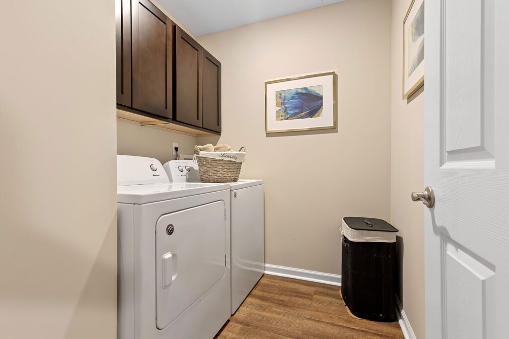 A laundry room with a washer and dryer, a basket on top, wall cabinets, framed artwork, a trash bin, and wood flooring.