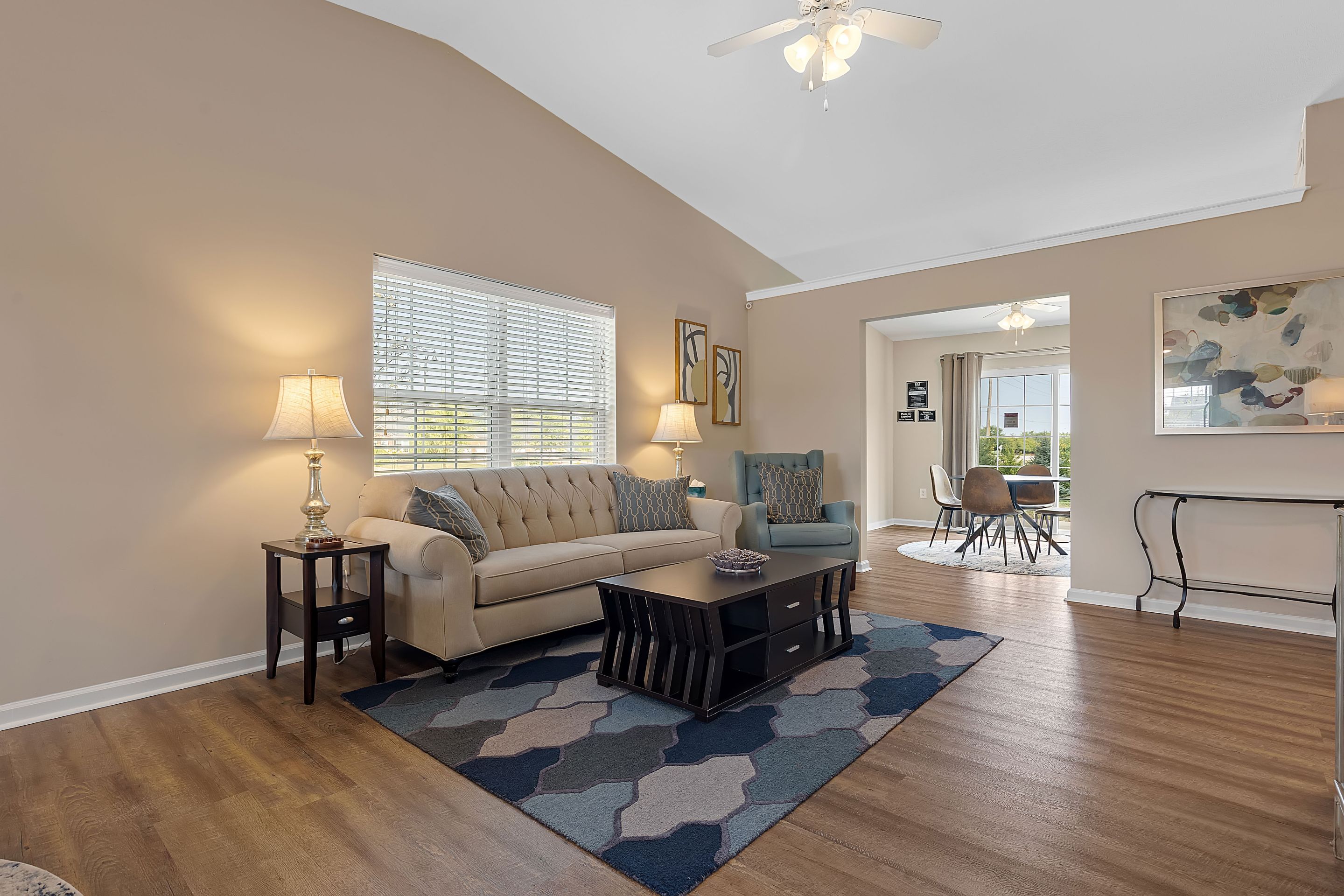 A living room with a beige sofa, armchair, coffee table, patterned rug, table lamps, and wall art, opening into a dining area with a round table and chairs.
