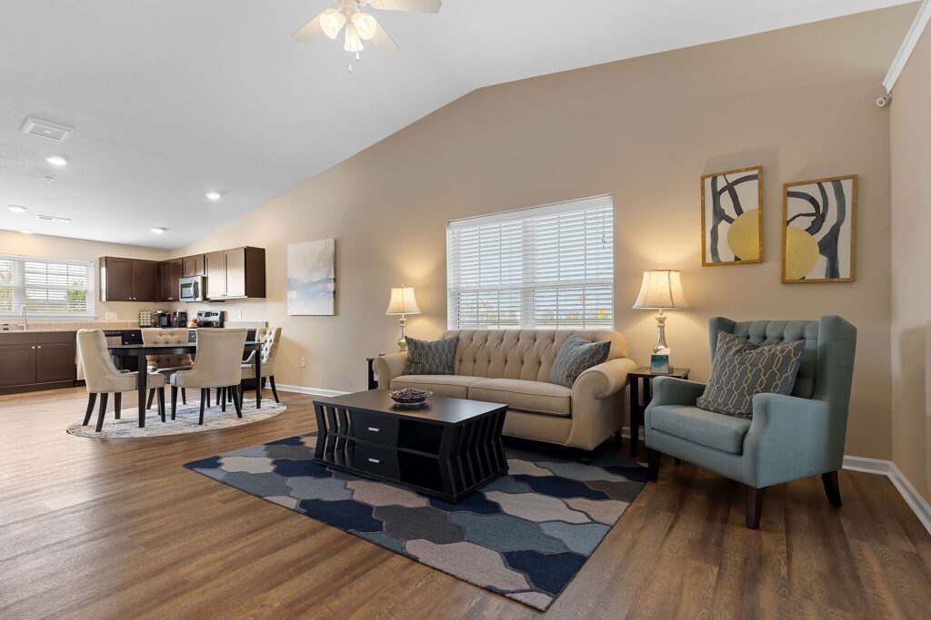 Open-plan living and dining area with a beige sofa, blue armchair, coffee table, lamps, wall art, and kitchen in the background. Hardwood floors and neutral walls throughout.