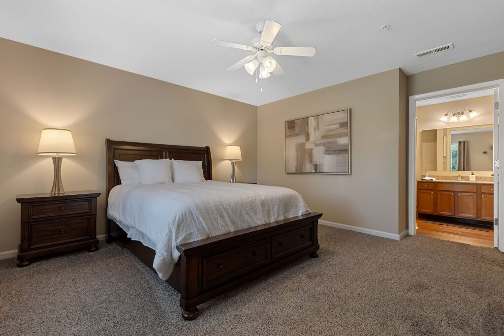 Bedroom with a wooden bed, two bedside tables with lamps, neutral walls, carpeted floor, artwork on the wall, and an open door leading to a bathroom.