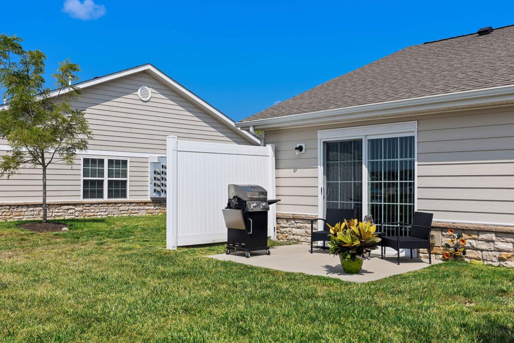 A backyard patio with a grill, two black chairs, a small table, and a potted plant, enclosed by a privacy fence next to a house with sliding glass doors.