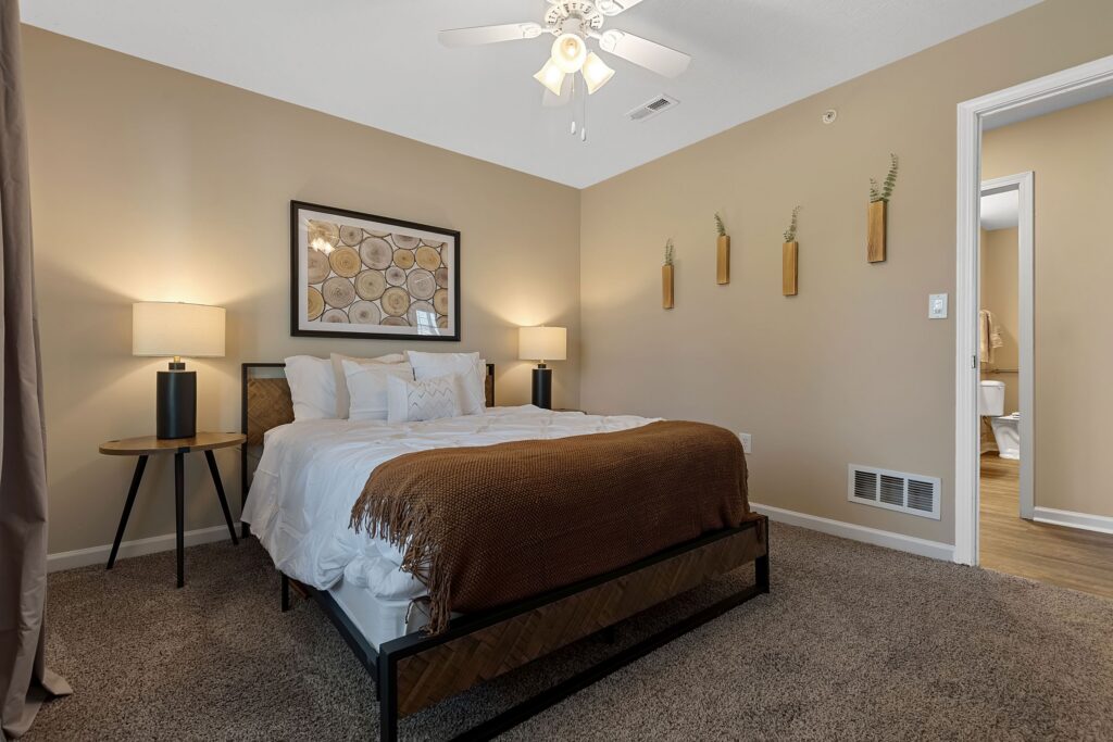 Bedroom with beige walls, bed with white bedding and brown blanket, two side tables with lamps, wall art above bed, and three decorative wall planters. Doorway leads to bathroom.
